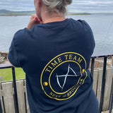 A woman viewed from behind wearing a navy blue t-shirt with a yellow and white circular graphic on the back that reads 'TIME TEAM' and 'NESS OF BRODGAR'. She is standing outdoors on a balcony overlooking a body of water under an overcast sky.