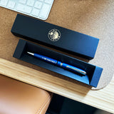 A blue ballpoint pen with silver trim and the text 'TIME TEAM IT IS BACK' resting inside an open black presentation box, with the matching closed box placed above it on a desk surface.