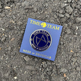 A circular enamel pin on a blue backing card, placed on a bed of dark gravel. The pin is dark blue with gold metal trim and bow or butterfly symbol, with the words 'TIME TEAM' and 'NESS OF BRODGAR'.